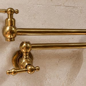 Puede incluir: Un grifo de llenado de ollas montado en la pared con un acabado de latón pulido. Este accesorio presenta un diseño de brazo oscilante articulado de doble articulación con dos manijas, una en la base y otra en el caño, ofreciendo alcance y flexibilidad.