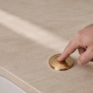 Pode incluir: Um tampão de drenagem circular de metal dourado com um design de borda chanfrada lisa. O tampão está embutido numa superfície de bancada de pedra bege clara texturizada, apresentando um mecanismo de botão circular central para operação.