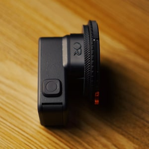 May include: A black action camera with a square body and a prominent circular lens housing featuring a textured grip ring. The side of the device displays a square power button and a small logo with the letters OR embossed on the lens mount.