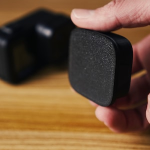 May include: A black foam wind reduction cover for an action camera, featuring a textured, porous surface. The rectangular accessory is designed to fit over the camera lens housing to minimise wind noise during outdoor recording sessions.