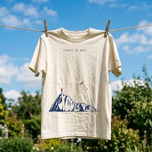 May include: A cream-coloured short-sleeve t-shirt featuring a dark blue graphic of a mountain peak with a person standing at the summit with arms raised. The text "Commit or Quit" is printed in a typewriter-style font above the mountain graphic.