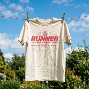 May include: A cream-colored short-sleeve t-shirt featuring red text that reads The Runner, Speed Meets Spirit, and Run Your Story Not The Race, with a small graphic of three running figures and the text Since 1991 printed on the front.