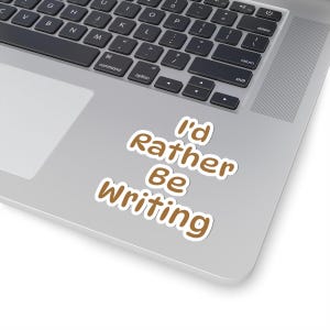 May include: A die-cut sticker with a white border featuring the brown text I'd Rather Be Writing in a rounded, bold font. The sticker is adhered to the silver metallic surface of a laptop near the black keyboard and trackpad area.