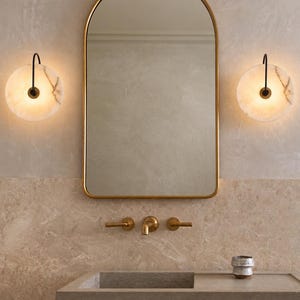 May include: An arched wall mirror with a thin gold frame, flanked by two circular wall sconces featuring white marble-like shades and black hardware. Below, a brass faucet set is mounted on a beige stone wall above a rectangular stone sink.
