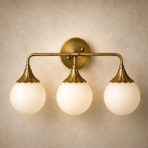 May include: A three-light wall sconce featuring a circular brass backplate and a horizontal bar supporting three downward-facing arms. Each arm holds a spherical, frosted white glass globe shade topped with a scalloped, antique brass cap.
