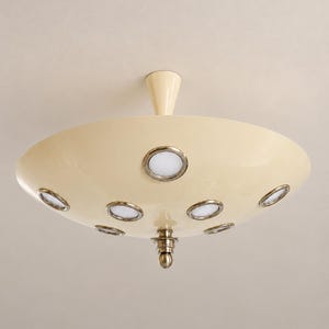May include: A cream-colored saucer-shaped ceiling light fixture featuring a central conical mount and a decorative metallic finial at the bottom. The surface is punctuated by multiple circular light ports with silver-toned rims and white glass.