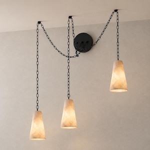 Puede incluir: Una lámpara colgante de tres luces con un soporte de pared circular negro y tres cadenas de metal negro. Cada cadena sostiene una pantalla cónica de piedra translúcida en color blanco roto que emite una luz cálida y suave.