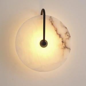 May include: A circular wall sconce featuring a translucent white stone disc with grey veining. A thin, matte black metal arm extends from the top, curving down to attach to the center of the disc, which emits a warm, soft glow from behind.