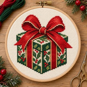 May include: A cross-stitch embroidery project in a circular wooden hoop featuring a square gift box wrapped in dark green paper with a holly leaf and berry pattern, topped with a large, vibrant red bow with gold-colored trim.
