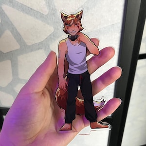 May include: A clear acrylic charm featuring a cartoon character with fox-like features. The character wears a white tank top, black pants, and a black collar. The charm is held in a hand, with a blurred background.