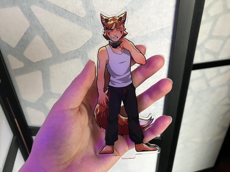 May include: A clear acrylic charm featuring a cartoon character with fox-like features. The character wears a white tank top, black pants, and a black collar. The charm is held in a hand, with a blurred background.