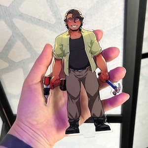 Op de afbeelding: Een heldere acrylfiguur van een man die een rood-zwarte boor en een hamer vasthoudt. De man draagt een groen overhemd over een zwart shirt, een bruine broek en zwarte laarzen. De figuur staat op een heldere basis.