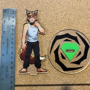 May include: An acrylic standee featuring a cartoon fox character in a white tank top and black pants, standing next to a circular base with a green diamond-shaped insert. A ruler shows the standee is approximately 6 inches tall.