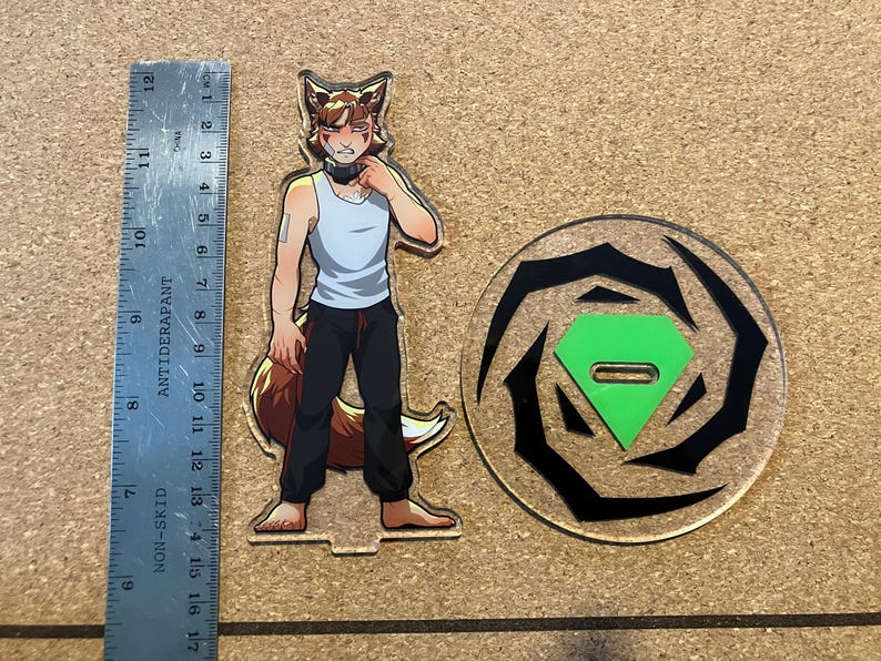 May include: An acrylic standee featuring a cartoon fox character in a white tank top and black pants, standing next to a circular base with a green diamond-shaped insert. A ruler shows the standee is approximately 6 inches tall.