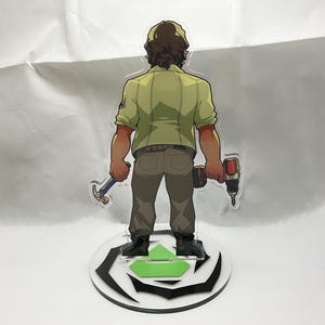 Op de afbeelding: Een heldere acrylfiguur van een persoon in een groen overhemd en bruine broek, met een hamer en een boor. De figuur staat op een cirkelvormige basis met een groen pijldesign en zwart-witte accenten.