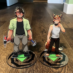 Op de afbeelding: Twee acryl standbeelden met stripfiguren. E&eacute;n houdt een boor en een hamer vast, draagt een groen overhemd, een zwart shirt en een grijze broek. De andere is een vosachtig personage in een witte tanktop en een zwarte broek. Beide staan op ronde bases.