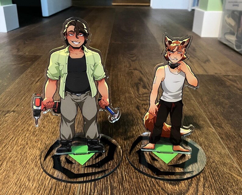 Op de afbeelding: Twee acryl standbeelden met stripfiguren. E&eacute;n houdt een boor en een hamer vast, draagt een groen overhemd, een zwart shirt en een grijze broek. De andere is een vosachtig personage in een witte tanktop en een zwarte broek. Beide staan op ronde bases.