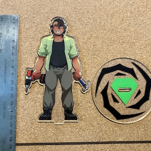Op de afbeelding: Een acrylfiguur van een lachende persoon met een boor en een hamer, met een bijpassende basis. De figuur draagt een groen overhemd over een zwart shirt, een grijze broek en zwarte laarzen. Een liniaal meet ongeveer 18 cm.