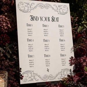 May include: A white rectangular wedding seating chart sign with black text and ornate scrollwork borders. The sign features the heading FIND YOUR SEAT followed by a list of nine tables, each containing the names Mary T, Steph L, Clara H, and Matt L.