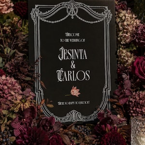 May include: A rectangular black wedding sign with white decorative border detailing and text reading Welcome to the wedding of Jesinta & Carlos, with a small floral illustration and the text We're so happy to have you printed below.