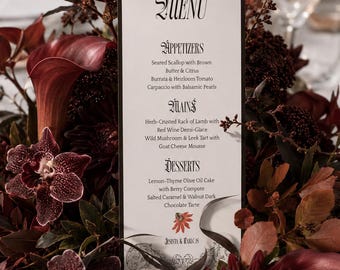 May include: A vertical white menu card with a black border and decorative scrollwork at the top and bottom. The text reads MENU, APPETIZERS, MAINS, DESSERTS, and lists various food items, ending with the names JESINTA & CARLOS and a small red flower.