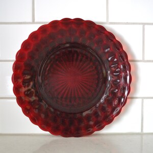 May include: A circular red glass plate featuring a scalloped edge and a textured hobnail pattern around the rim. The center of the plate displays a radiating sunburst design with fine, linear grooves extending outward from the middle.