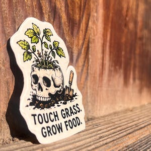 May include: A die-cut sticker featuring a human skull acting as a planter for a green leafy plant. A small brown shovel is stuck in the soil next to the skull. The text "TOUCH GRASS. GROW FOOD." is printed in black capital letters at the bottom.