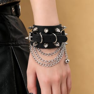 May include: A black leather cuff bracelet featuring silver-toned metal spikes arranged in two rows. The design includes an adjustable buckle strap, two circular metal rings, and three dangling silver-toned chain layers with a small spherical charm.