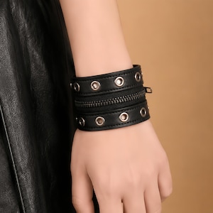 May include: A black cuff bracelet featuring two horizontal bands of smooth material, each accented with a row of silver-toned metal grommets. A central horizontal zip runs between the two bands, adding a textured detail to the design.