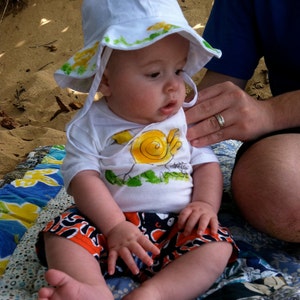 Op de afbeelding: Een jong kind draagt ​​een witte zonnehoed met gele en groene bloemenmotieven en zit op een kleurrijk patchworkdeken. Het kind draagt ​​een wit T-shirt met een geel en groen slakkenmotief en oranje en zwarte shorts.