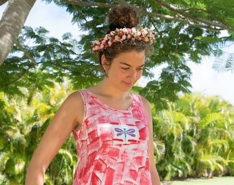 May include: A woman wearing a pink and white patterned tank top with a dragonfly design. She is wearing a flower crown and white pants.
