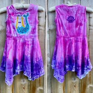 K&ouml;nnte beinhalten: Ein pinkfarbenes und violettes, tie-dye-farbenes, &auml;rmelloses Kleid mit einem Meerjungfrauen-Motiv auf der Vorderseite und einem Muschel-Motiv auf der R&uuml;ckseite. Das Kleid hat einen R&uuml;schen-Saum und ist aus einem weichen, bequemen Stoff gefertigt.