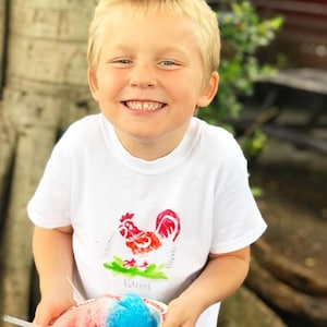 May include: A young boy wearing a white t-shirt with a red rooster design and the word "Kauai" printed on it. He is holding a bowl of shaved ice with red and blue flavors.