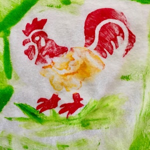 Op de afbeelding: Een witte stof met een rode, gele en groene haan erop geschilderd. De haan staat op een groen veld.