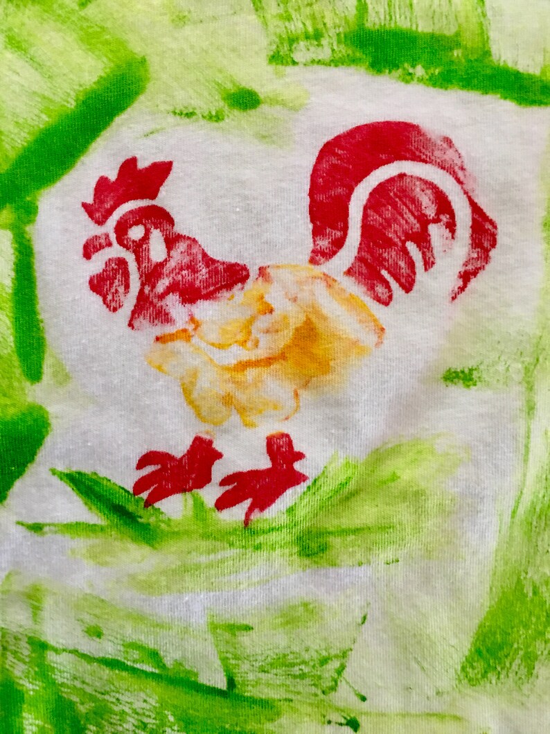 Op de afbeelding: Een witte stof met een rode, gele en groene haan erop geschilderd. De haan staat op een groen veld.