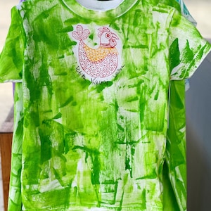 Op de afbeelding: Groen T-shirt met een wit kippenontwerp en de tekst "Eat me, Kanuri, will you?"