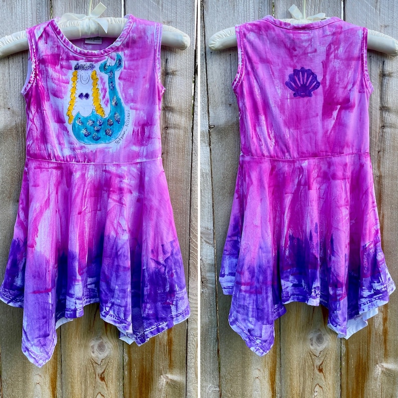 K&ouml;nnte beinhalten: Ein pinkfarbenes und violettes, tie-dye-farbenes, &auml;rmelloses Kleid mit einem Meerjungfrauen-Motiv auf der Vorderseite und einem Muschel-Motiv auf der R&uuml;ckseite. Das Kleid hat einen R&uuml;schen-Saum und ist aus einem weichen, bequemen Stoff gefertigt.