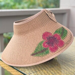 Puede incluir: Una visera de sol de paja color canela con un ala ancha y curva y un dise&ntilde;o abierto en la parte superior. El frente de la visera est&aacute; decorado con un motivo floral pintado que consiste en una flor rosa vibrante con centros morados oscuros.