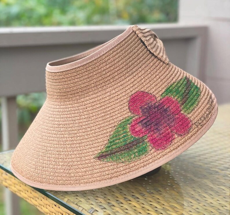 Puede incluir: Una visera de sol de paja color canela con un ala ancha y curva y un dise&ntilde;o abierto en la parte superior. El frente de la visera est&aacute; decorado con un motivo floral pintado que consiste en una flor rosa vibrante con centros morados oscuros.