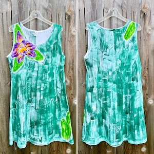 Peut inclure: Une robe sans manches avec un motif abstrait de coups de pinceau verts peints &agrave; la main. Le devant pr&eacute;sente une grande fleur d'orchid&eacute;e violette et jaune avec des feuilles vertes, tandis que le dos inclut un d&eacute;tail de feuille verte pr&egrave;s de l'&eacute;paule.