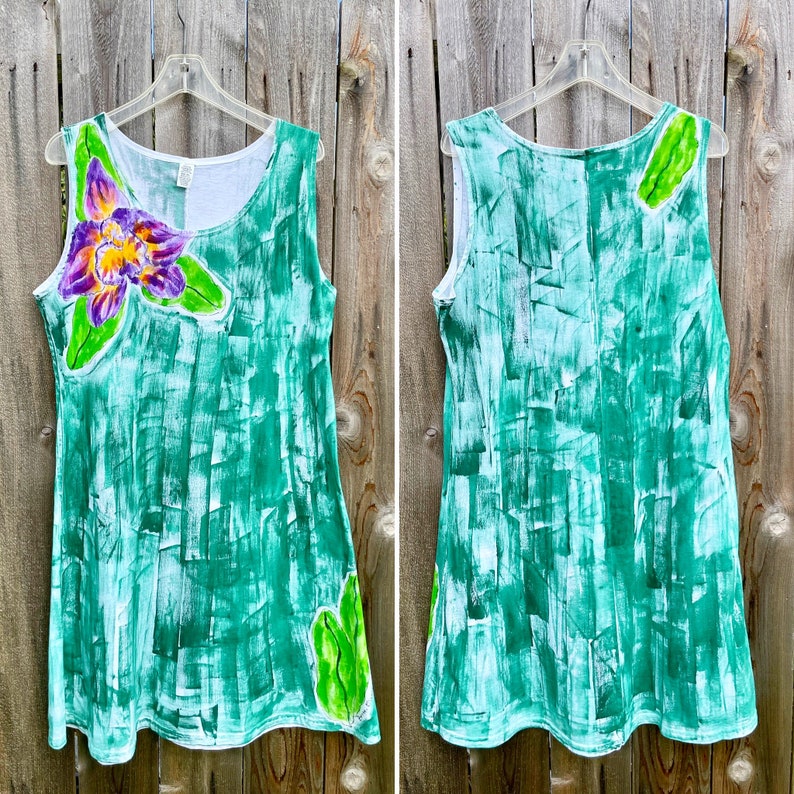 Peut inclure: Une robe sans manches avec un motif abstrait de coups de pinceau verts peints &agrave; la main. Le devant pr&eacute;sente une grande fleur d'orchid&eacute;e violette et jaune avec des feuilles vertes, tandis que le dos inclut un d&eacute;tail de feuille verte pr&egrave;s de l'&eacute;paule.