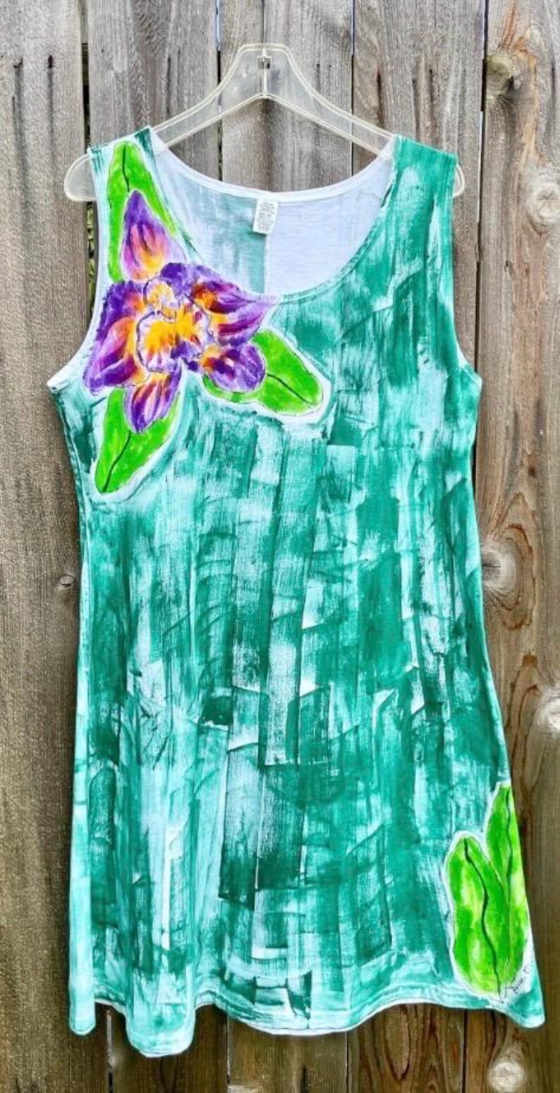 Peut inclure: Une robe sans manches avec un motif abstrait peint &agrave; la main en coups de pinceau verts. Le v&ecirc;tement pr&eacute;sente une grande fleur d'orchid&eacute;e violette et jaune sur le haut de la poitrine gauche et des feuilles vertes sur l'ourlet inf&eacute;rieur droit.