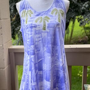 Puede incluir: Una camiseta sin mangas con base blanca y un patr&oacute;n de bloques estampados en estilo acuarela azul. La zona superior del pecho muestra tres palmeras verdes impresas. La prenda tiene un cuello redondo y un dobladillo asim&eacute;trico.