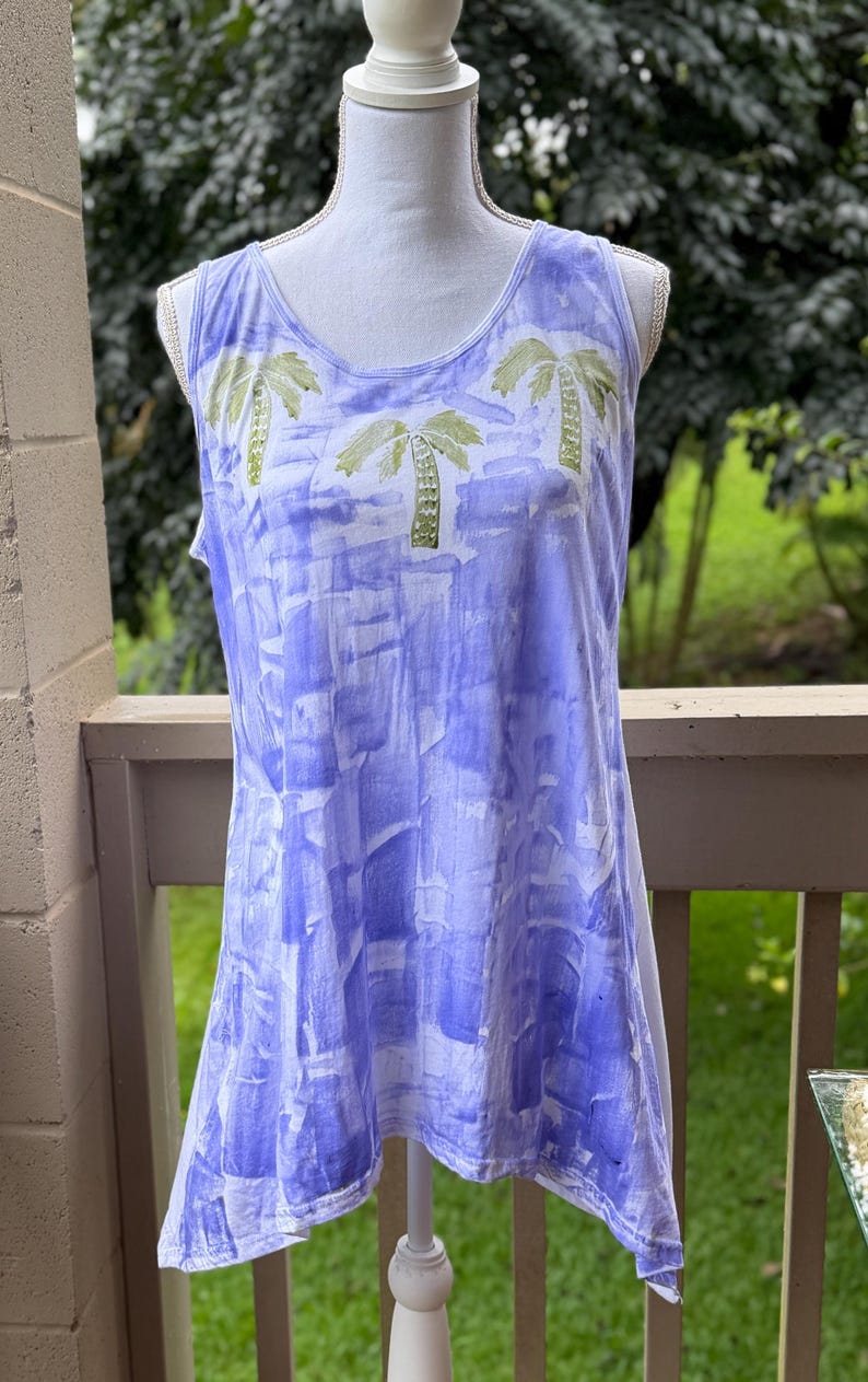 Puede incluir: Una camiseta sin mangas con base blanca y un patr&oacute;n de bloques estampados en estilo acuarela azul. La zona superior del pecho muestra tres palmeras verdes impresas. La prenda tiene un cuello redondo y un dobladillo asim&eacute;trico.