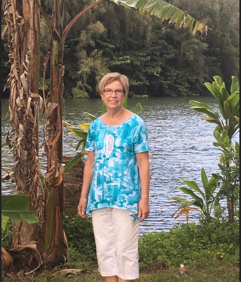 May include: A short-sleeved tunic top featuring a vibrant blue and white abstract pattern with a central white feather graphic, paired with solid white cropped pants. The top has a rounded neckline and a slightly asymmetrical hemline.