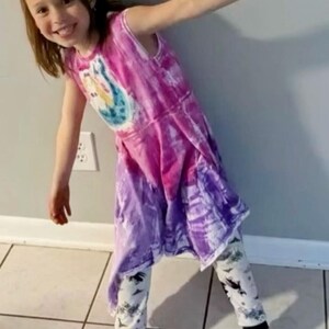 K&ouml;nnte beinhalten: Ein junges Kind tr&auml;gt ein Batik-Kleid in Lila und Rosa. Das Kleid hat ein grafisches Design auf der Vorderseite. Das Kind tr&auml;gt auch schwarze und wei&szlig;e gemusterte Leggings.