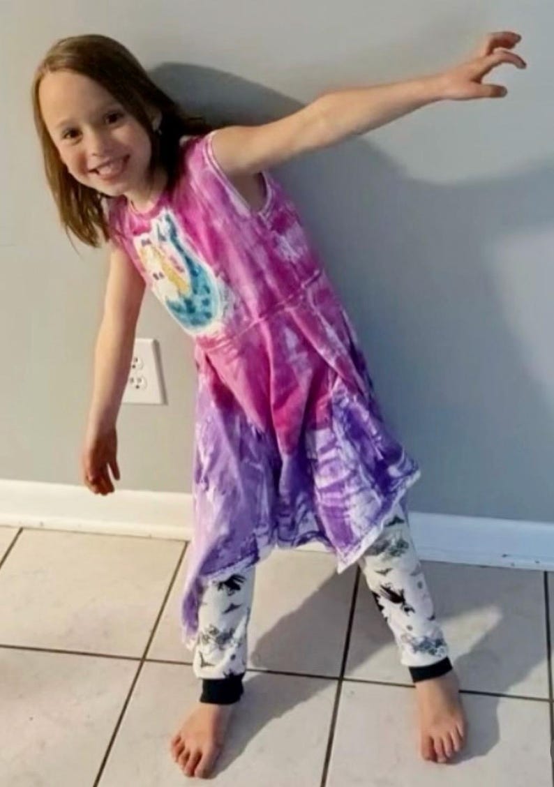 K&ouml;nnte beinhalten: Ein junges Kind tr&auml;gt ein Batik-Kleid in Lila und Rosa. Das Kleid hat ein grafisches Design auf der Vorderseite. Das Kind tr&auml;gt auch schwarze und wei&szlig;e gemusterte Leggings.