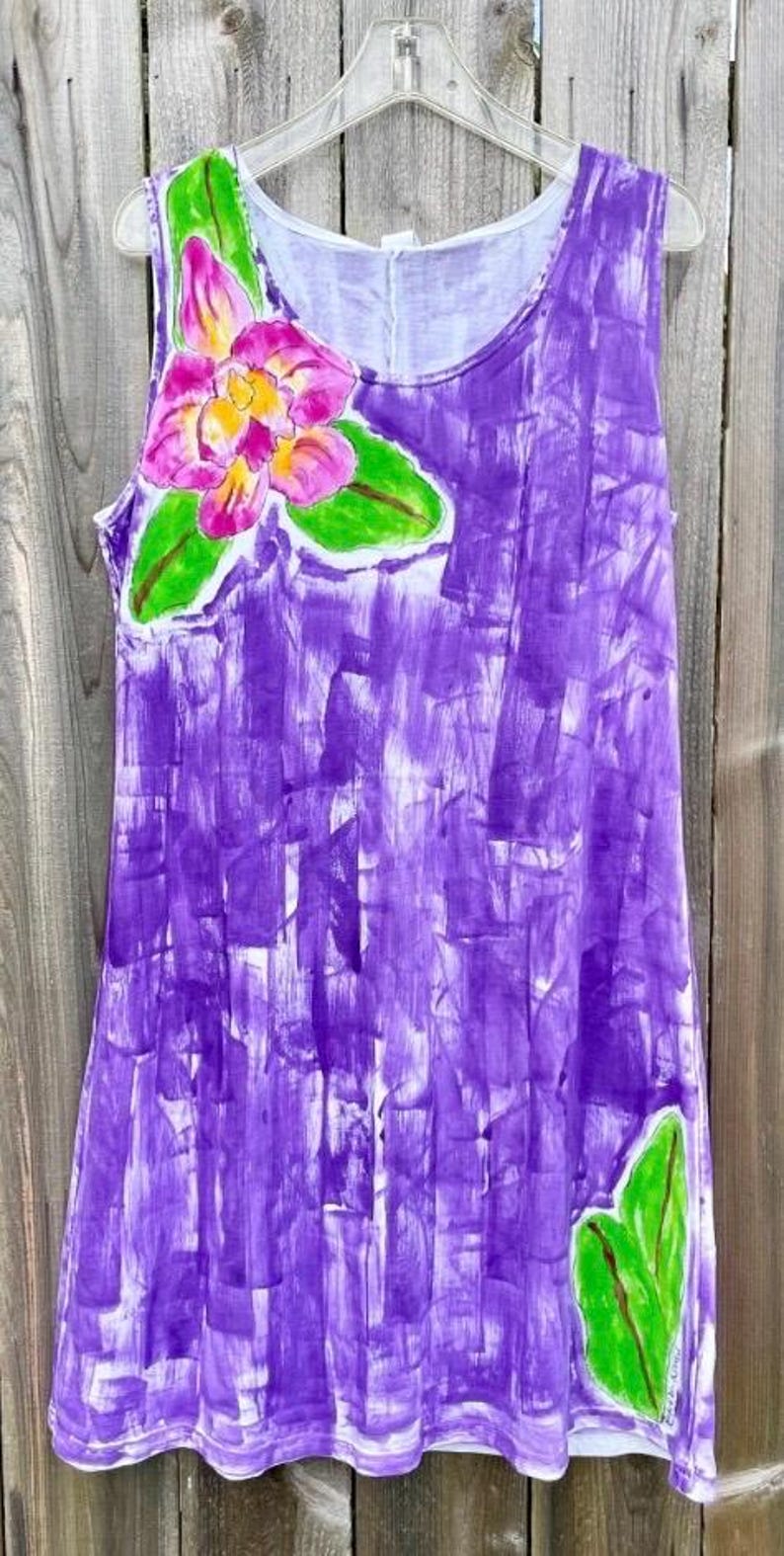 Peut inclure: Une robe sans manches avec un motif abstrait peint &agrave; la main en coups de pinceau violets. Le v&ecirc;tement pr&eacute;sente une grande fleur rose et jaune avec des feuilles vertes pr&egrave;s de l'encolure et une feuille verte pr&egrave;s de l'ourlet inf&eacute;rieur.