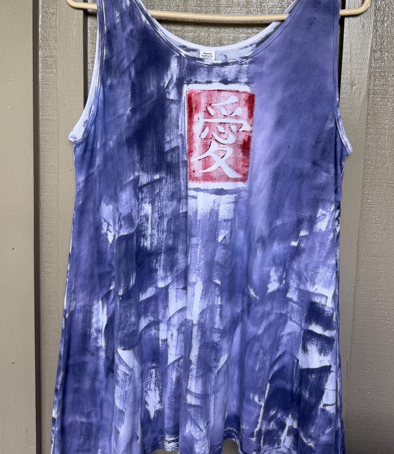 May include: A sleeveless tank top featuring a purple and white tie-dye pattern with a rectangular graphic on the chest. The graphic contains a red background with the white Japanese kanji character for love, which is written as 愛, centered on the fabric.