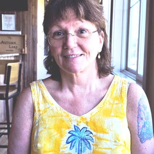 May include: A sleeveless yellow and white tie-dye top featuring a blue palm tree graphic on the chest. The garment has a V-neckline and a relaxed fit. The person is wearing thin-rimmed glasses and small gold drop earrings with a visible arm tattoo.